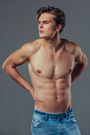 Handsome young man isolated. Shirtless muscular man in jeans is standing on grey background. Man holding his back. Experiencing backache.の写真素材