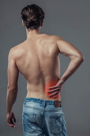 Handsome young man isolated. Shirtless muscular man in jeans is standing on grey background. Man holding his back. Experiencing backache.の写真素材