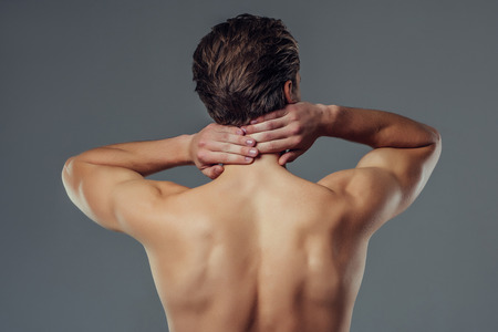 Handsome young man isolated. Shirtless muscular man is standing on grey background. Back view of man holding his neck. Experiencing neck pain.の写真素材