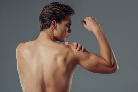 Handsome young man isolated. Shirtless muscular man is standing on grey background. Back view of man holding his shoulder. Experiencing shoulder pain.の写真素材
