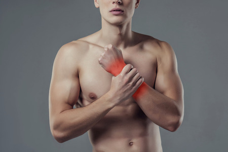 Handsome young man isolated. Cropped image of shirtless muscular man is standing on grey background. Close-up of man holding his wrist. Experiencing wrist pain.の写真素材