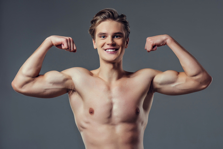 Handsome young man isolated. Portrait of shirtless muscular man is standing on grey background, showing his biceps, smiling and looking at camera.の写真素材