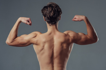 Handsome young man isolated. Portrait of shirtless muscular man is standing on grey background and showing his biceps. Back viewの写真素材