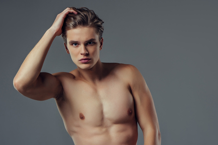 Handsome young man isolated. Portrait of shirtless muscular man is standing on grey background and holding his hair.の写真素材