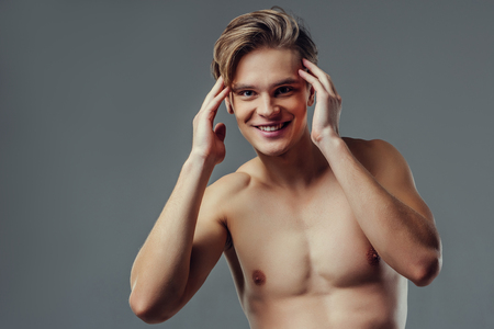 Handsome young man isolated. Portrait of shirtless muscular man is standing on grey background and holding his hair.の写真素材