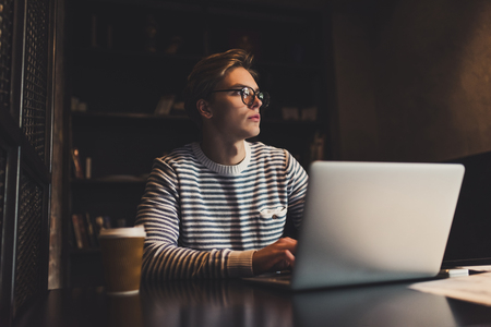 Young handsome man in eyeglasses is drinking coffee while working in cafe with laptop.の写真素材