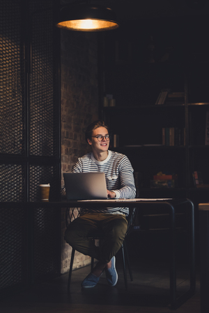 Young handsome man in eyeglasses is drinking coffee while working in cafe with laptop.の写真素材