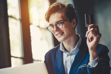 Young handsome stylish businessman in eyeglasses is working in cafe with laptop. Holding credit card in hand and smiling.の写真素材