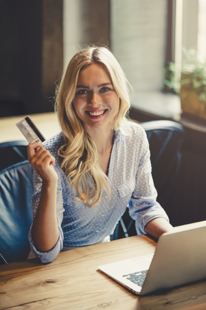 Young attractive businesswoman is working in cafe with laptop. Holding credit card in hand, looking at camera and smiling.の写真素材