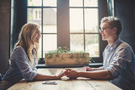 Handsome young man and attractive young woman are spending time together. Romantic couple in cafe is drinking coffee, holding hands and enjoying being together.の写真素材
