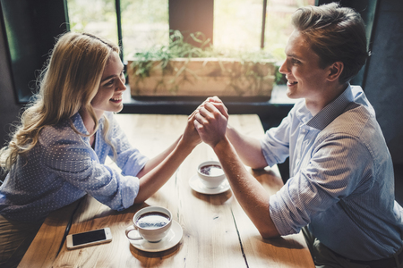 Handsome young man and attractive young woman are spending time together. Romantic couple in cafe is drinking coffee, holding hands and enjoying being together.の写真素材