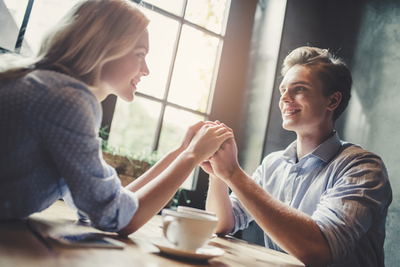 Handsome young man and attractive young woman are spending time together. Romantic couple in cafe is drinking coffee, holding hands and enjoying being together.の写真素材