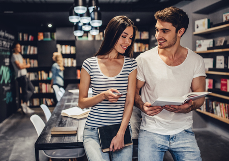 Students are studying in library. Young people are spending time together. Young handsome men and attractive women are working together. Holding mobile phone and books in hands.の写真素材