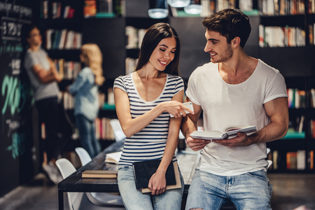 Students are studying in library. Young people are spending time together. Young handsome men and attractive women are working together. Holding mobile phone and books in hands.の写真素材