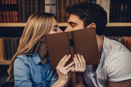 Students are studying in library. Young people are spending time together. Romantic couple is kissing while reading book.の写真素材