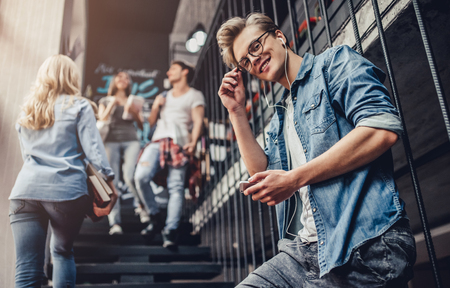 Students are studying in library. Young people are spending time together. Young handsome man in earrings is listening to music with smart phone while standing on stairs.の写真素材