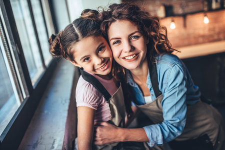 Attractive young woman and her little cute daughter are cooking on kitchen. Having fun together while making cakes and cookies. Hugging, smiling and looking at camera.の写真素材