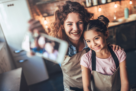 Attractive young woman and her little cute daughter are cooking on kitchen. Having fun together while making cakes and cookies. Smiling and making selfie on smart phone.の写真素材