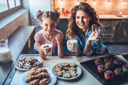 Attractive young woman and her little cute daughter are eating cakes and cookies on kitchen and drinking milk. Having fun together while enjoying 
freshly baked pastries. Smiling and looking at camera.の写真素材