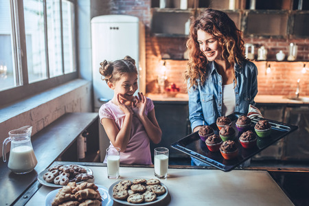 Attractive young woman and her little cute daughter are eating cakes and cookies on kitchen and drinking milk. Having fun together while enjoying 
freshly baked pastries.の写真素材