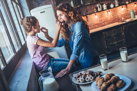 Attractive young woman and her little cute daughter are eating cakes and cookies on kitchen and drinking milk. Having fun together while enjoying 
freshly baked pastries.の写真素材