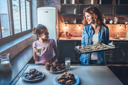 Attractive young woman and her little cute daughter are eating cakes and cookies on kitchen and drinking milk. Having fun together while enjoying 
freshly baked pastries.の写真素材