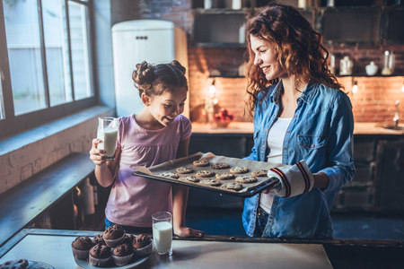 Attractive young woman and her little cute daughter are eating cakes and cookies on kitchen and drinking milk. Having fun together while enjoying 
freshly baked pastries.の写真素材