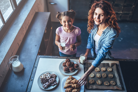 Top view of attractive young woman and her little cute daughter are eating cakes and cookies on kitchen and drinking milk. Having fun together while enjoying 
freshly baked pastries.の写真素材