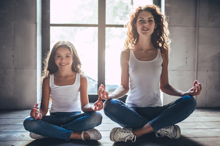 Beautiful curly woman and her little pretty daughter at home. Practicing yoga in lotus posture.の写真素材
