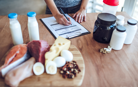Hard choice. Cropped image of young sporty woman is choosing between healthy food full of protein and sport supplements while standing on light kitchen and making notes. Sports nutrition.の写真素材