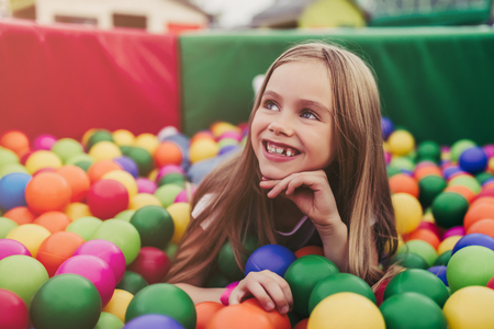 Little pretty girl is having fun outdoor. Playing in children zone in amusement park. Lying in colorful balls and smilingの写真素材