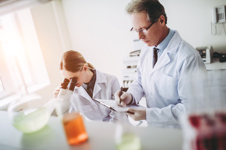 Two scientists are working in laboratory. Young female researcher and her senior supervisor are doing investigations with microscope and test tubes.の写真素材