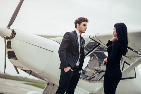 Beautiful young stylish couple  in official clothes is standing near private plane. Looking eye to eye in airport in front of airplane.の写真素材