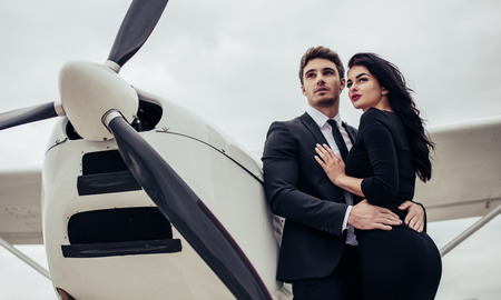 Beautiful young stylish couple in official clothes is standing near private plane. Hugging and kissing in airport in front of airplane.の写真素材