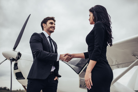 Beautiful young stylish couple  in official clothes is standing near private plane. Shaking hands in airport in front of airplane.の写真素材