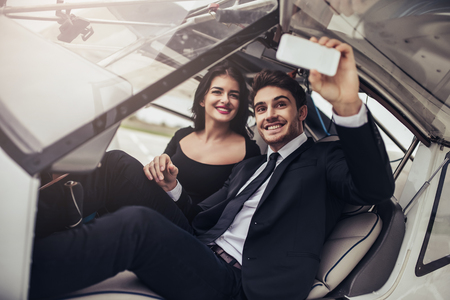 Beautiful young romantic couple in official clothes is sitting in private plane. Smiling and making selfie on smart phone.の写真素材