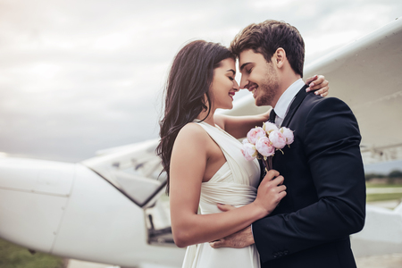 Just married! Beautiful young romantic couple is hugging and kissing near private plane. Attractive woman in wedding dress and handsome man in suit are celebrating wedding day in airport near airplane. Ready for Honeymoon.の写真素材