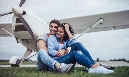 Beautiful romantic couple is sitting on grass near private plane in airport. Handsome man and attractive woman are hugging and smiling.の写真素材