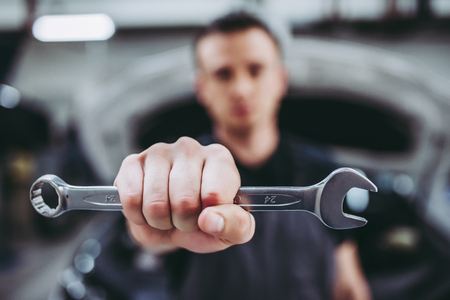 Handsome auto service mechanic in uniform is standing on the background of car with open hood with wrench in hand. Car repair and maintenance.の写真素材