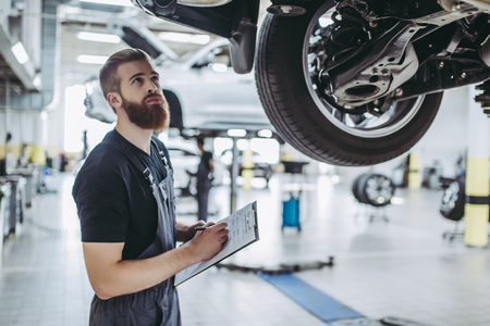 Handsome mechanic in uniform is examining the car while working in auto service with lifted vehicle. Car repair and maintenance.の写真素材