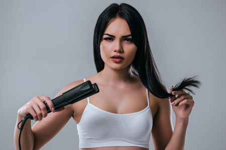 Portrait of attractive young woman with hair straightener holding the tips of hair on grey background. Women careの写真素材