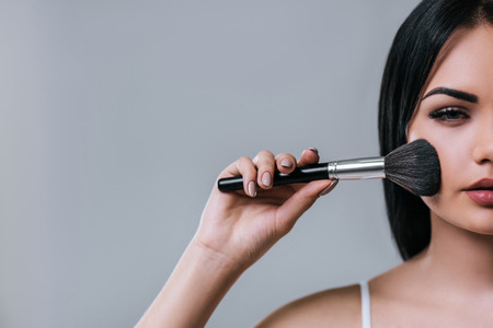 Cropped image of attractive young woman with makeup brush on grey background. Half faceの写真素材