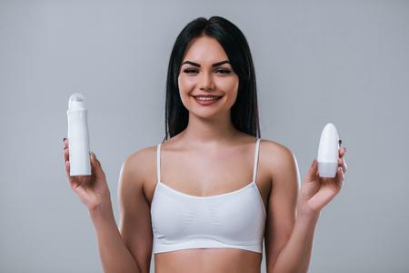 Portrait of attractive young woman with two deodorants on grey background. Making choice. Women careの写真素材