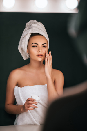 Attractive young woman in bathroom after shower is standing in front of mirror with face cream. Women care.の写真素材