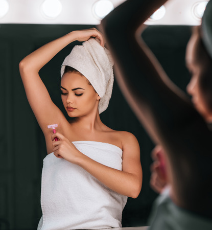 Attractive young woman in bathroom after shower is standing in front of mirror with razor in hand while shaving. Women care.の写真素材