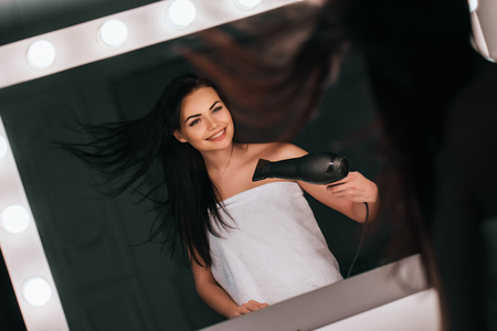Attractive young woman in bathroom after shower is standing in front of mirror with hair dryer in hand. Women care.の写真素材