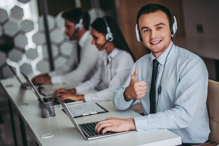 How can I help you? Beautiful call center workers in headphones are working at modern office. Handsome man is smiling and showing thumb up.の写真素材
