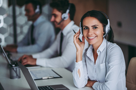 How can I help you? Beautiful call center workers in headphones are working at modern office.の写真素材