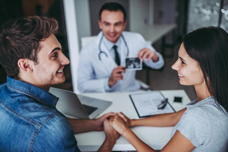 Young couple at the doctor. Holding hands and looking on each other while doctor is showing the ultrasond picture of their future baby.の写真素材