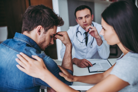 Young couple at the doctor. Attractive woman is calming her man. Everything is gonna be okay.の写真素材